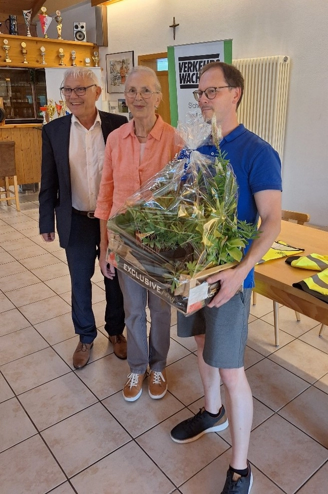 Ehrung bei einer Veranstaltung der Verkehrswacht: Drei Personen präsentieren einen Geschenkkorb mit Pflanzen in einem Vereinsheim. Im Hintergrund sind ein Verkehrswacht-Banner und Auszeichnungen zu sehen.