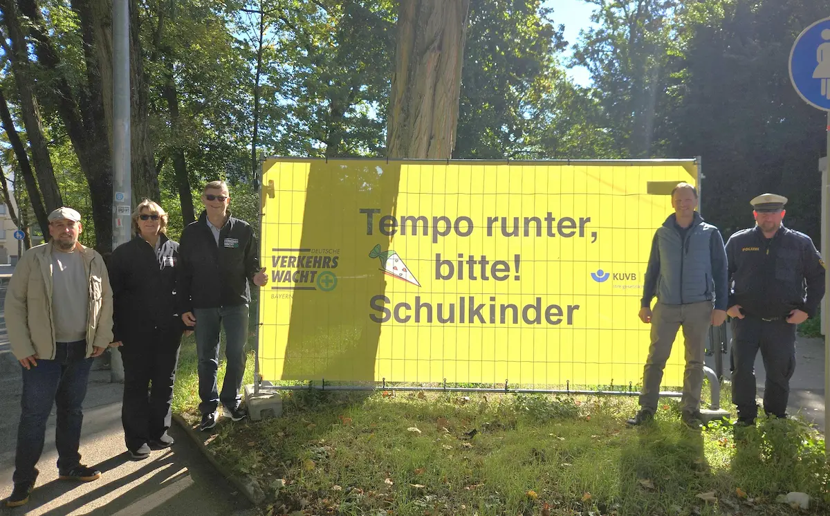 Banner der Verkehrssicherheitskampagne „Tempo runter, bitte! Schulkinder“ an einer Straße vor einem Schulweg. Mehrere Beteiligte stehen neben dem gelben Hinweisschild, das Autofahrende zu mehr Rücksicht und langsamerem Fahren im Bereich von Schulkindern auffordert.